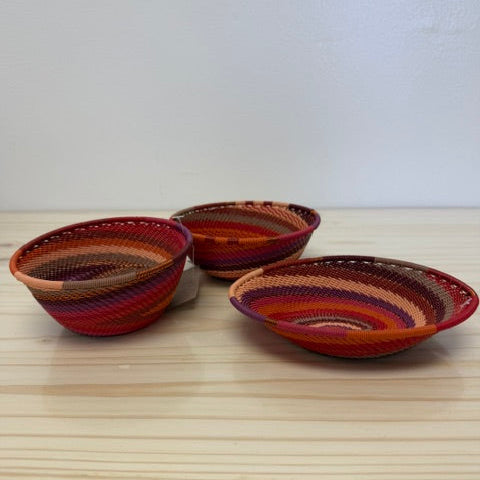 Three small hand-woven zulu baskets with colorful red, orange, brown, and purple stripes, arranged on a light wood tabletop against a plain white wall.