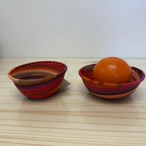 Two small handwoven zulu baskets in red, orange, and brown tones, with one basket holding an orange fruit, set on a light wood surface against a plain white wall.