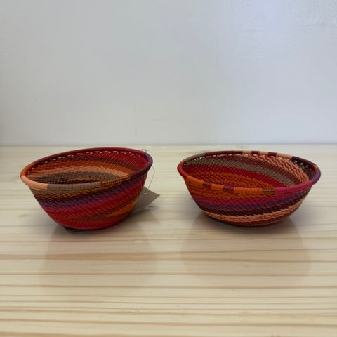 Two small handwoven zulu baskets with red, orange, and brown striped patterns, displayed on a light wood surface against a plain white wall.