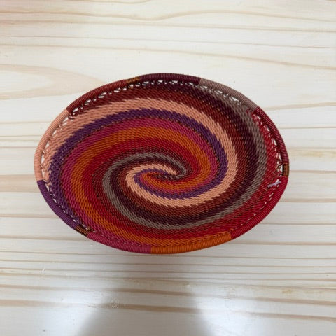 A small oval woven zulu basket with a spiral pattern in red, orange, brown, purple, and beige, placed on a light wood surface.
