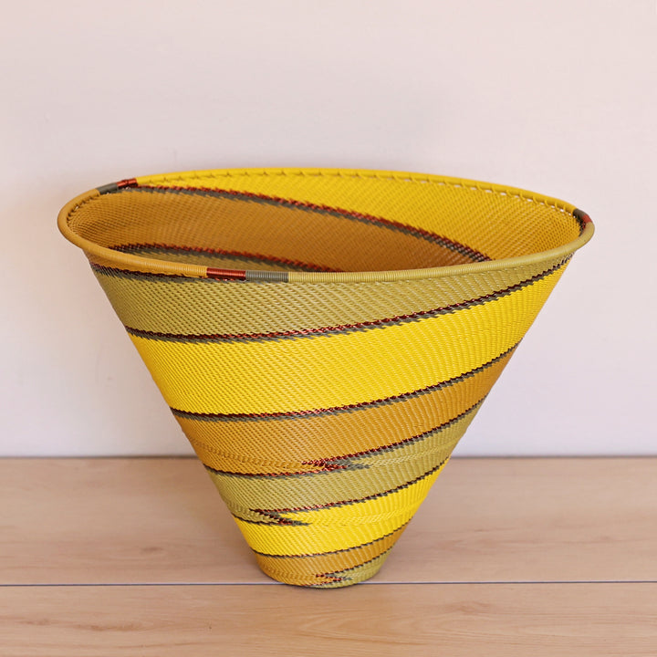 A papaya large deep funnel bowl basket made from yellow and olive-green striped material, displayed on a light wood surface against a plain off-white background.