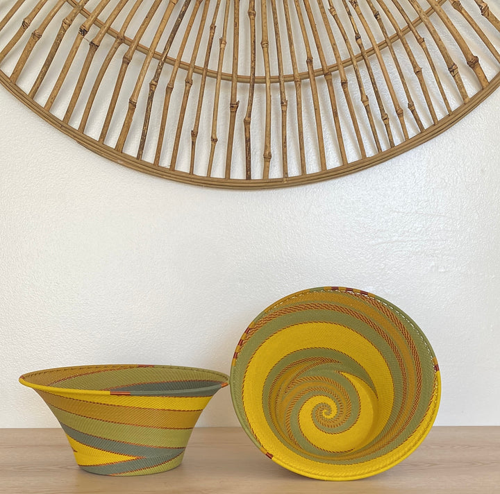 Two handwoven papaya large flared zulu decorative bowls displayed on a light wood surface against a white wall, with a large circular wooden wall hanging above. One bowl is shown from the side and the other from the top, both featuring yellow, olive green, and gray woven patterns.