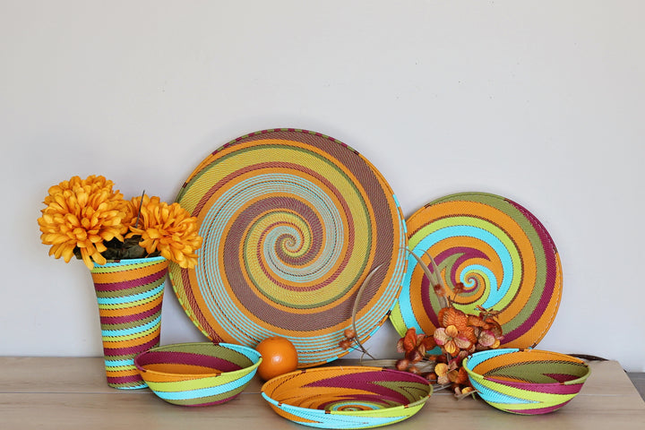A set of pastel Zulu handwoven decorative tableware with vibrant spiral patterns in orange, yellow, green, blue, and purple, arranged on a light wood table against a white wall with orange flowers in a striped vase.