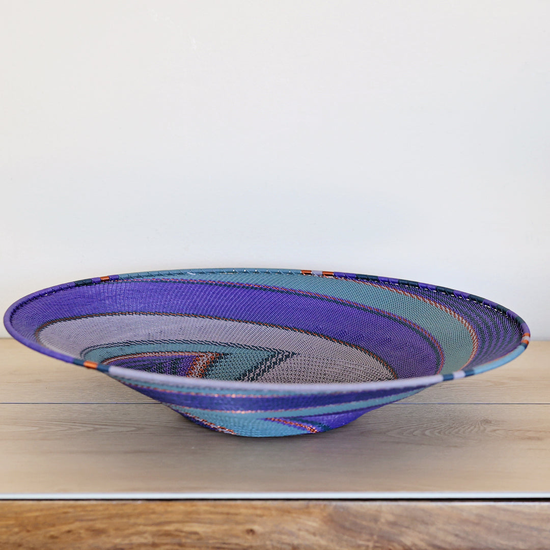 A hand-woven zulu basket bowl with concentric patterns of purple, teal, gray, and orange, displayed on a light wood surface against a white background.