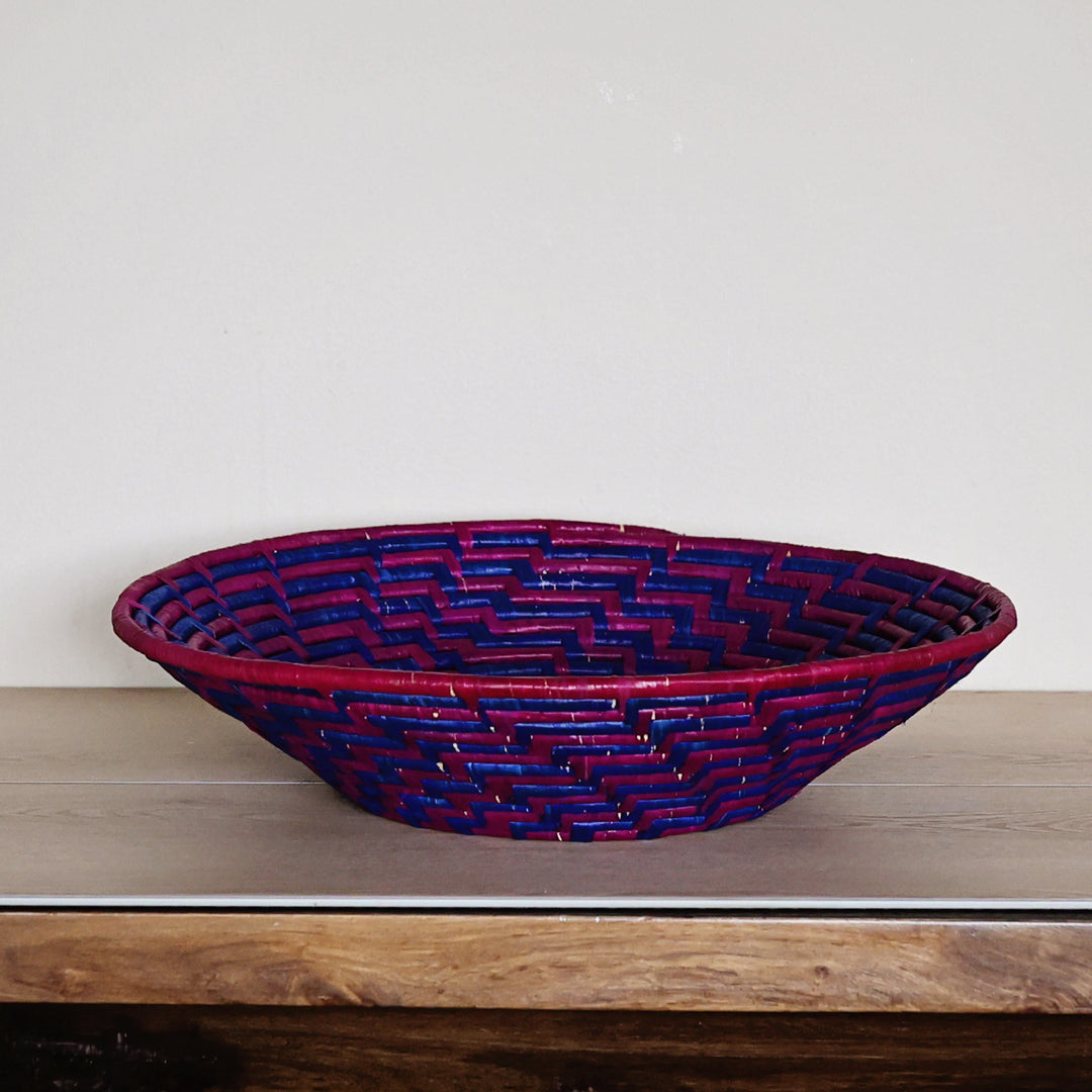 Handwoven purple and blue raffia fruit basket featuring an intricate woven pattern and shallow round silhouette.