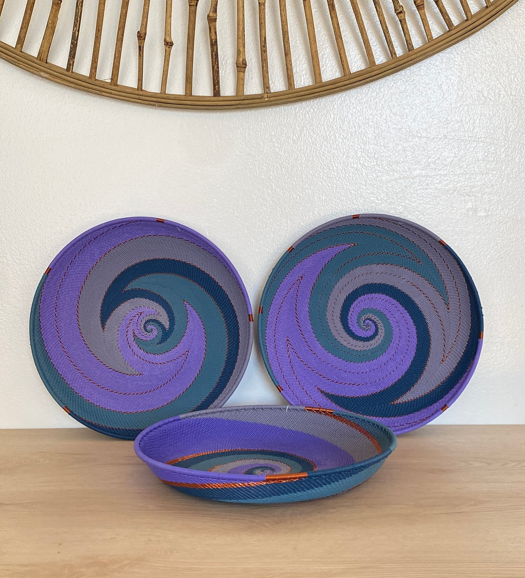 Three handwoven purple zulu large tray decorative baskets with purple, blue, and gray spiral patterns, displayed on a light wood surface against a white wall.