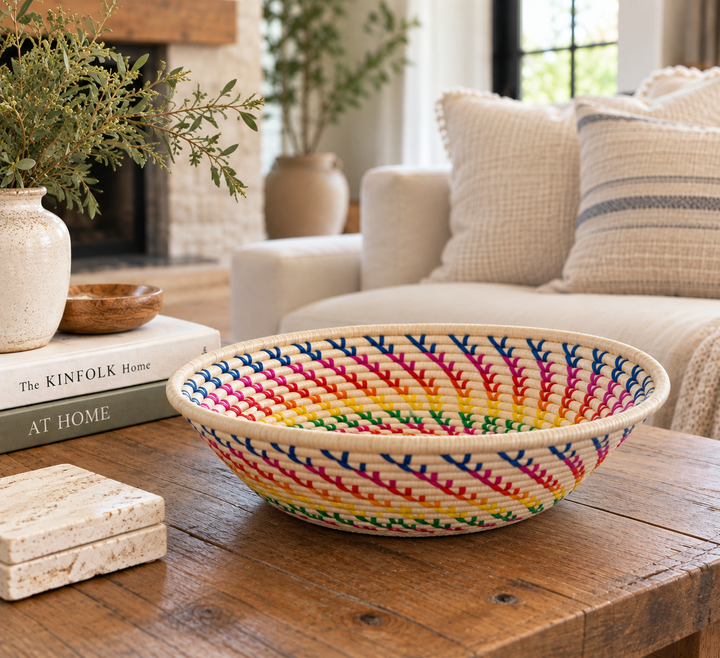 A round woven decorative rainbow raffia bowl with a colorful leaf-like pattern in pink, blue, yellow, and green, set on a rustic wooden table in a cozy living room.