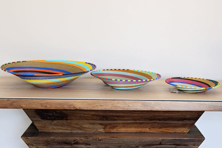 Three hand-woven striped Zulu  bowls in different sizes displayed on a rustic wooden table against a plain off-white wall.
