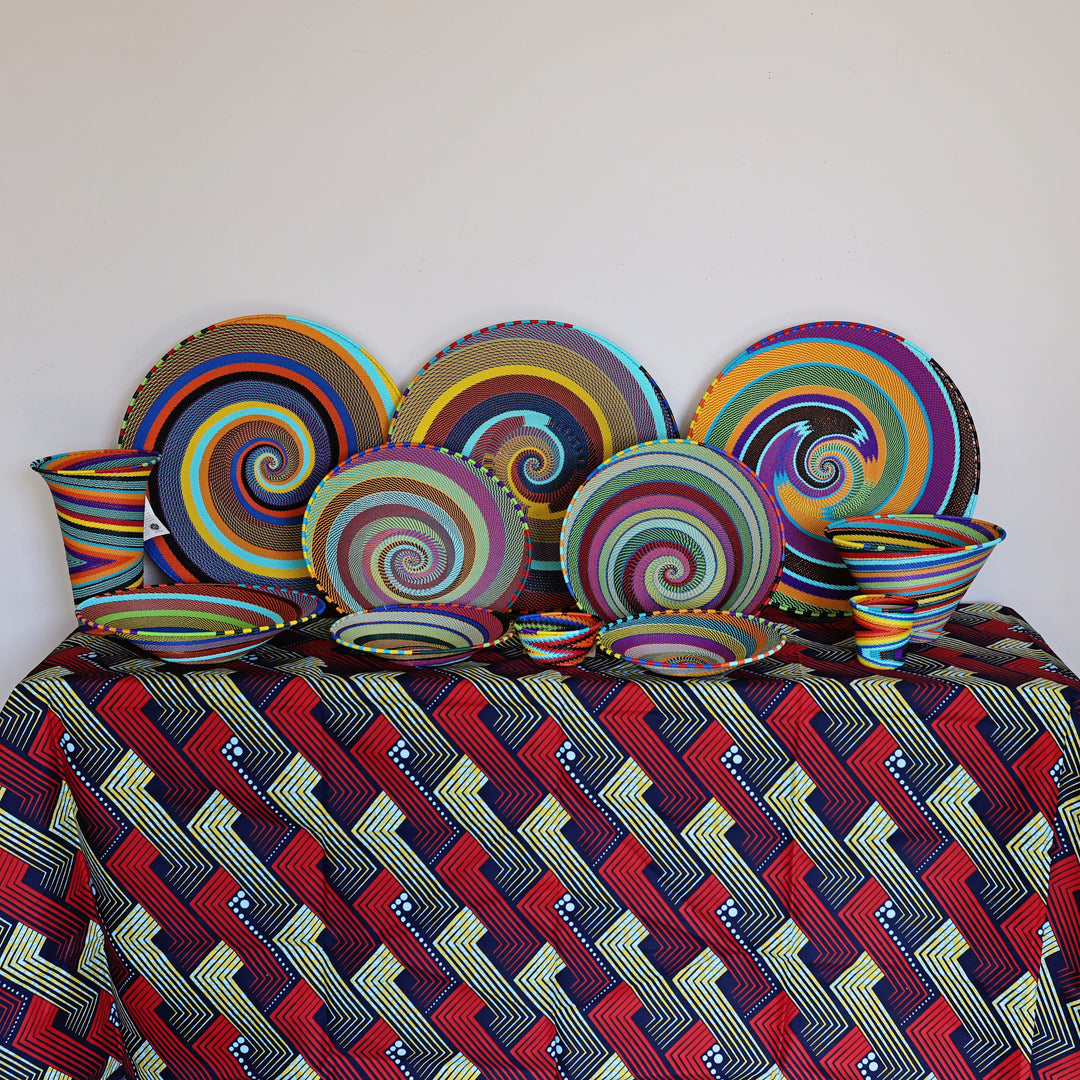 A colorful zulu tableware set arranged on a patterned tablecloth against a plain white wall. The set includes spiral-patterned plates, bowls, and a matching cup, with a geometric patterned tablecloth underneath.