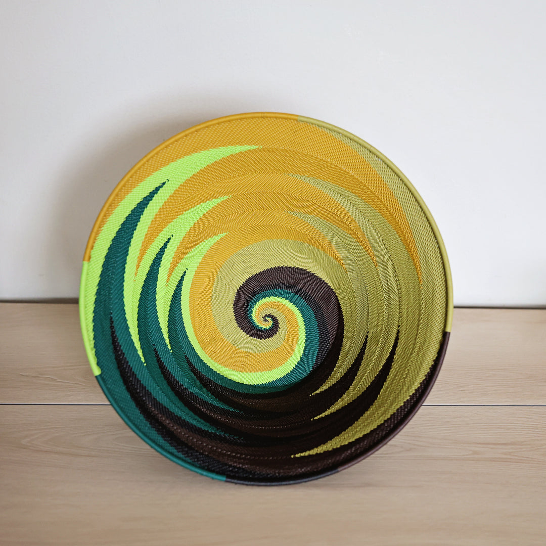 A round woven basket with a vibrant spiral pattern in yellow, green, teal, and dark brown, set against a light wood floor and white wall background.