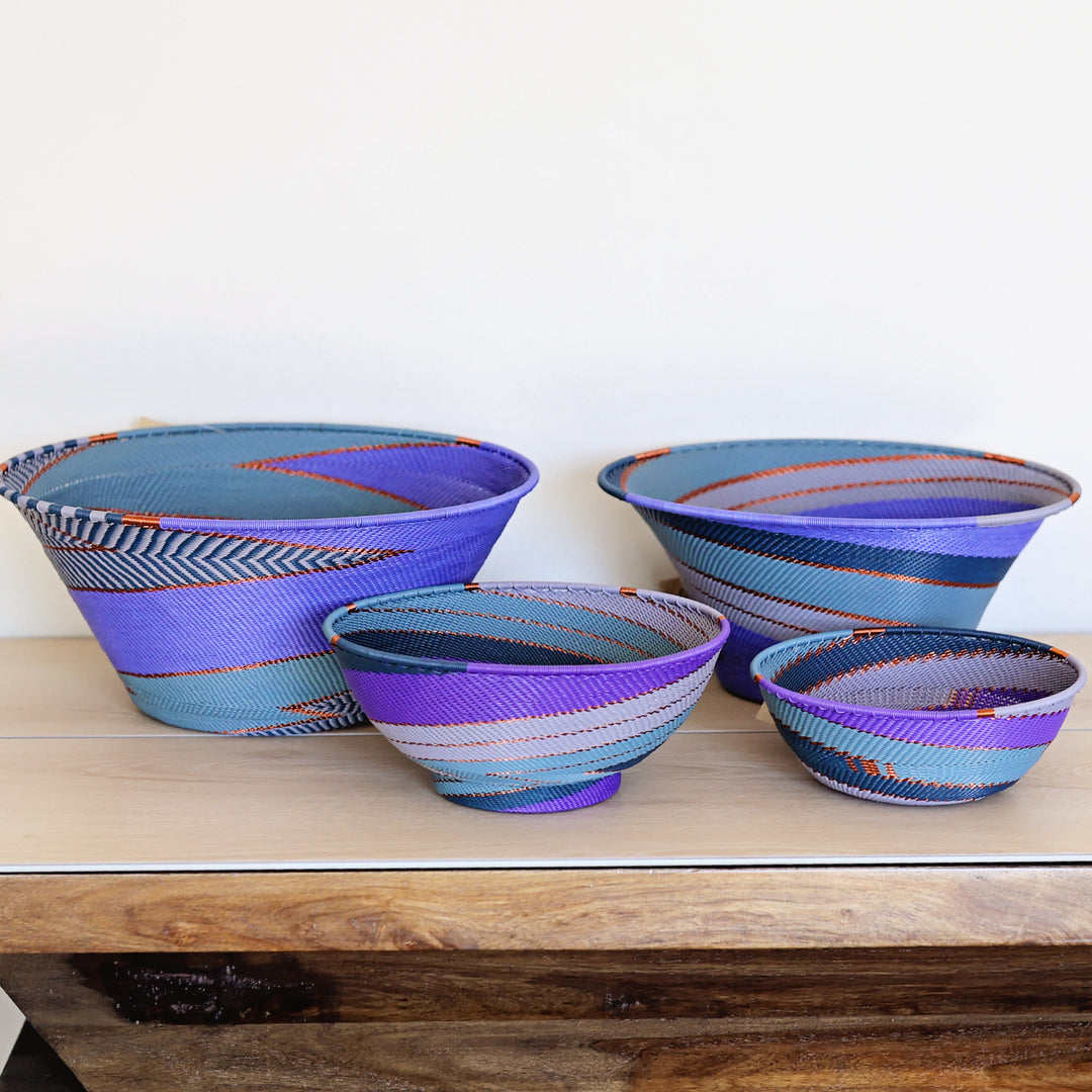 Four hand-woven zulu basket bowls in various sizes, decorated with vibrant purple, blue, and teal patterns, arranged on a light wood surface against a plain white wall.