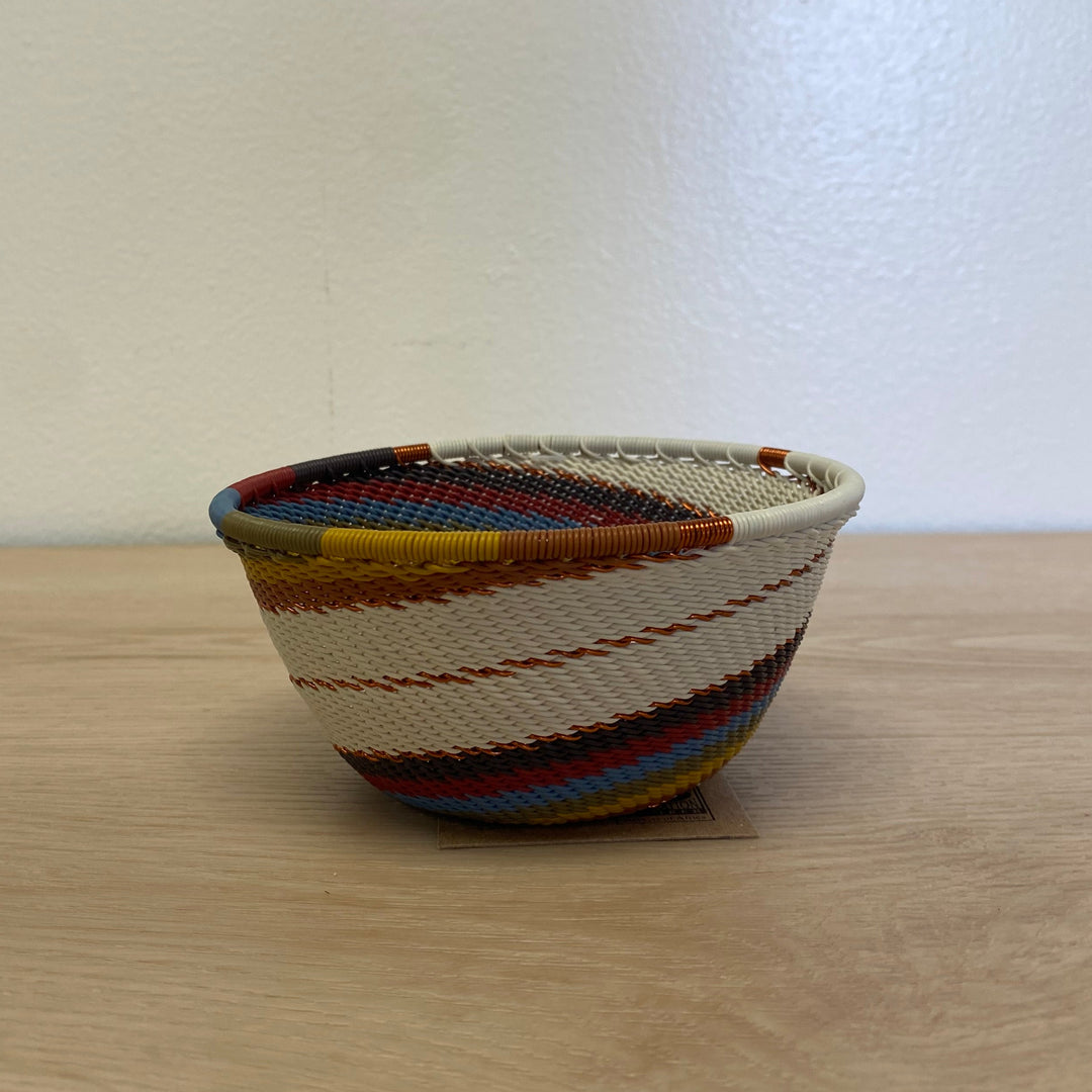 A small hand-woven mountain rainbow zulu basket bowl with multicolored stripes, placed on a light wood tabletop against a plain off-white wall.