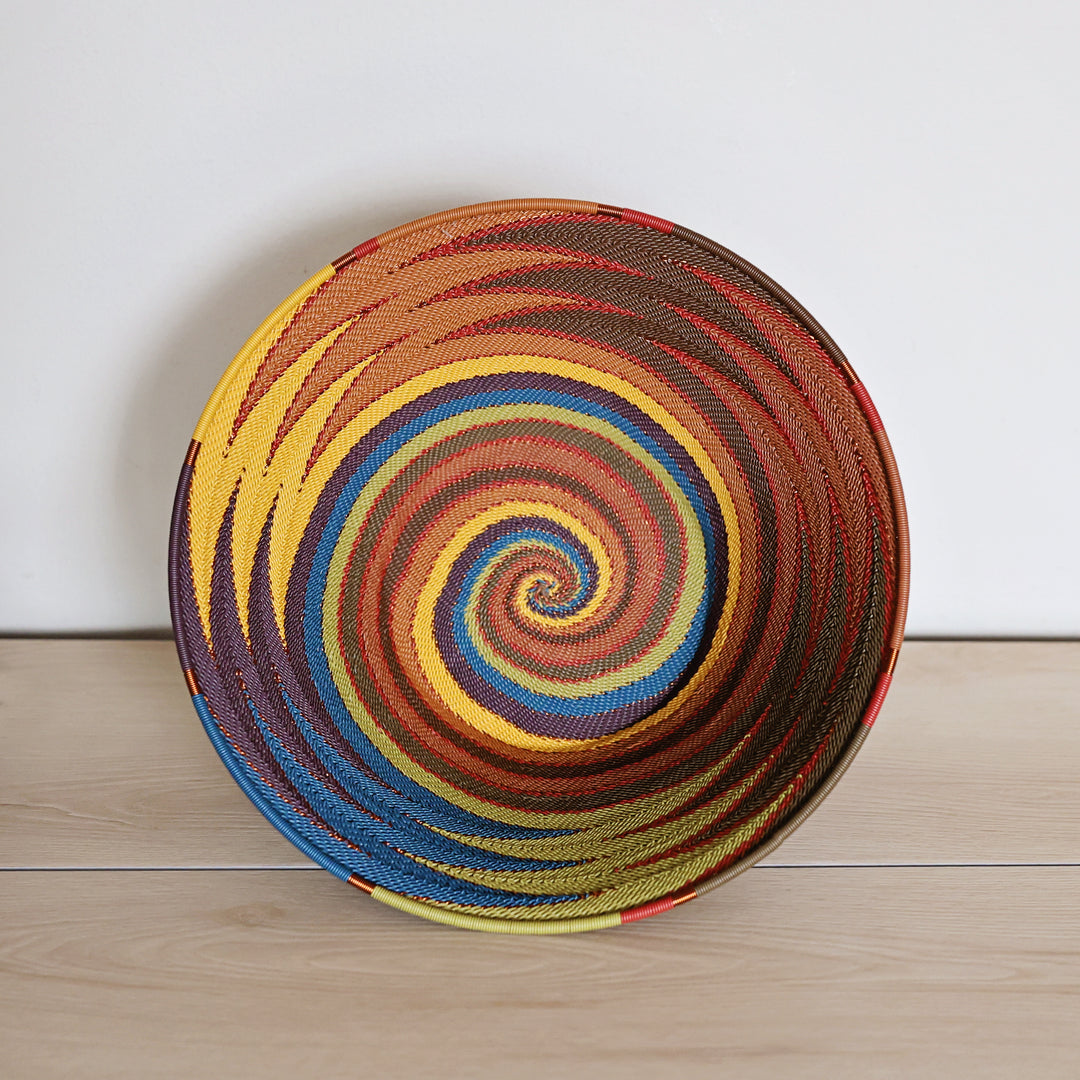 A round handwoven basket with a vibrant spiral pattern in yellow, orange, red, blue, and brown, set against a plain white wall and light wood floor.