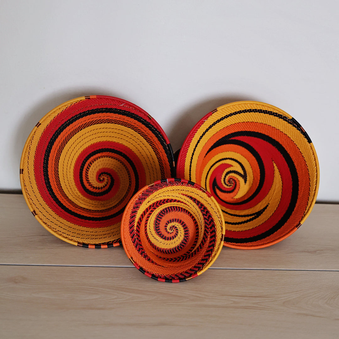 Three hand-woven spiral zulu baskets in vibrant red, orange, yellow, and black, arranged on a light wood surface against a plain white wall.
