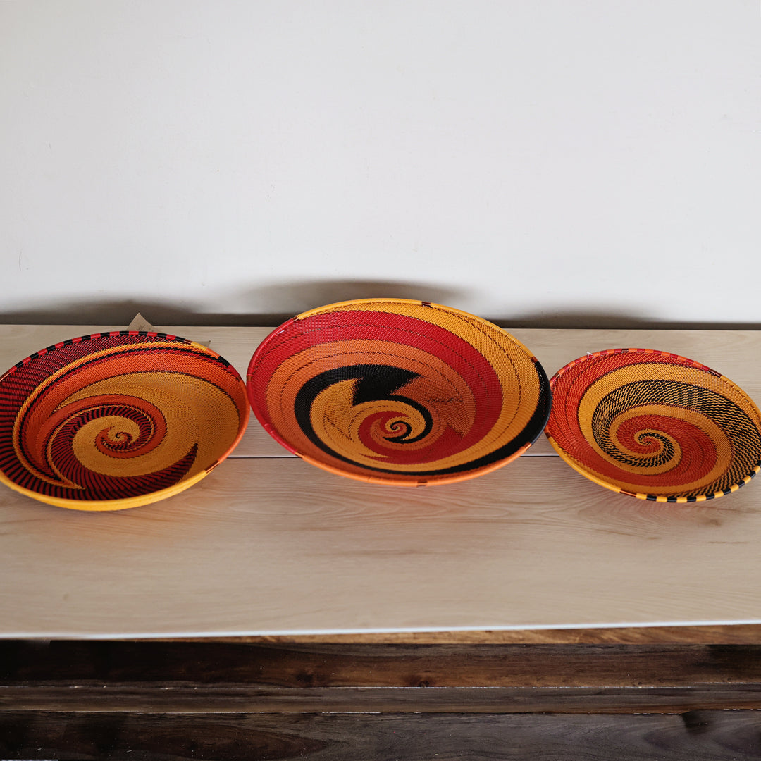 Three hand-woven decorative sunset zulu baskets with vibrant spiral patterns in red, orange, yellow, and black, arranged on a light-colored wooden shelf against a plain white wall.