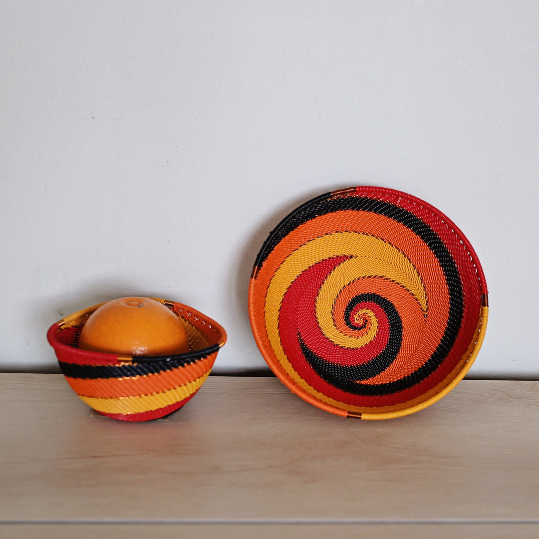Two hand-woven African zulu baskets with vibrant spiral patterns in red, orange, yellow, and black, displayed on a light wood surface against a plain white wall.