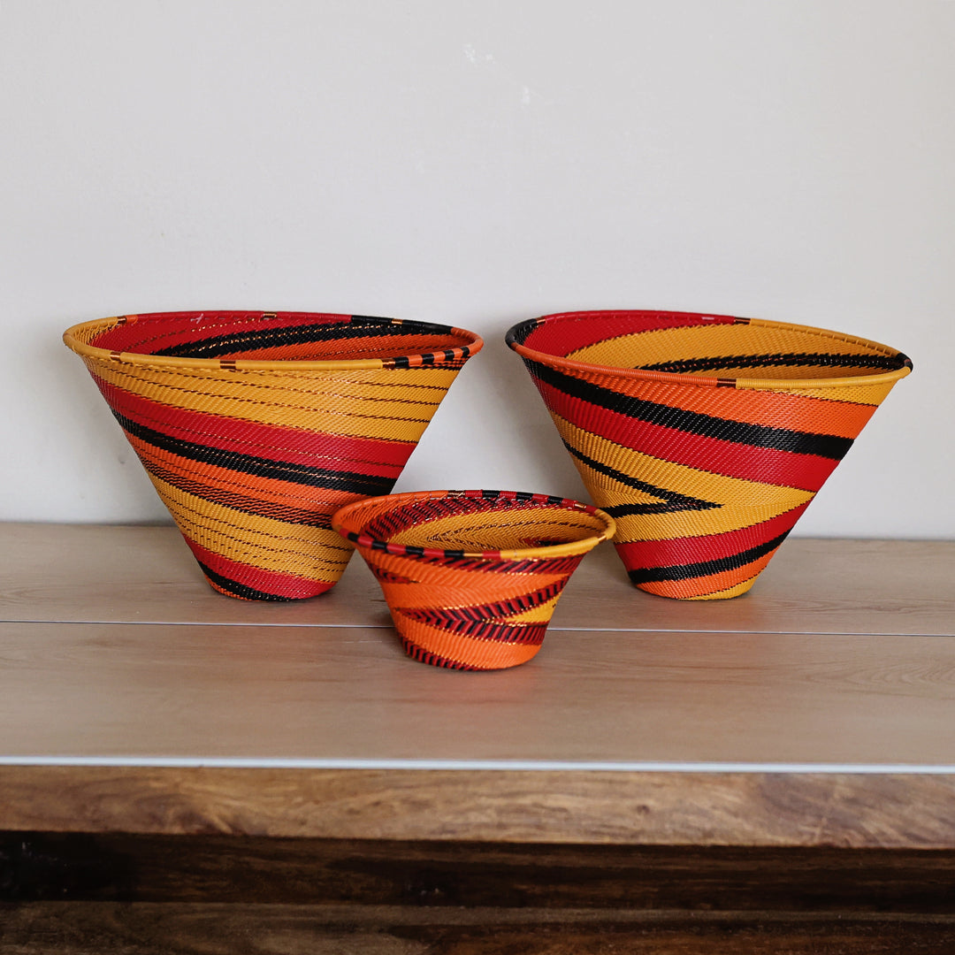 Three hand-woven baskets in different sizes with vibrant orange, yellow, red, and black stripes, arranged on a wooden shelf against a plain white wall.