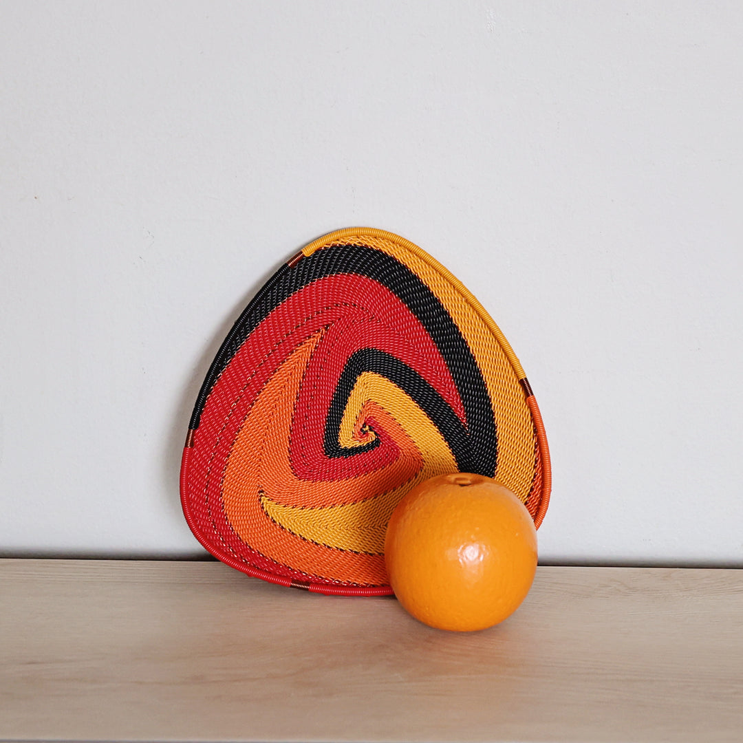 A triangular woven decorative zulu mat with a spiral pattern in red, orange, yellow, and black, leaning against a white wall on a light wood surface with an orange in front.