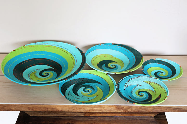 Five hand-woven decorative bowls with colorful spiral patterns in turquoise, lime green, black, and blue, arranged on a light wood table against a plain white wall.