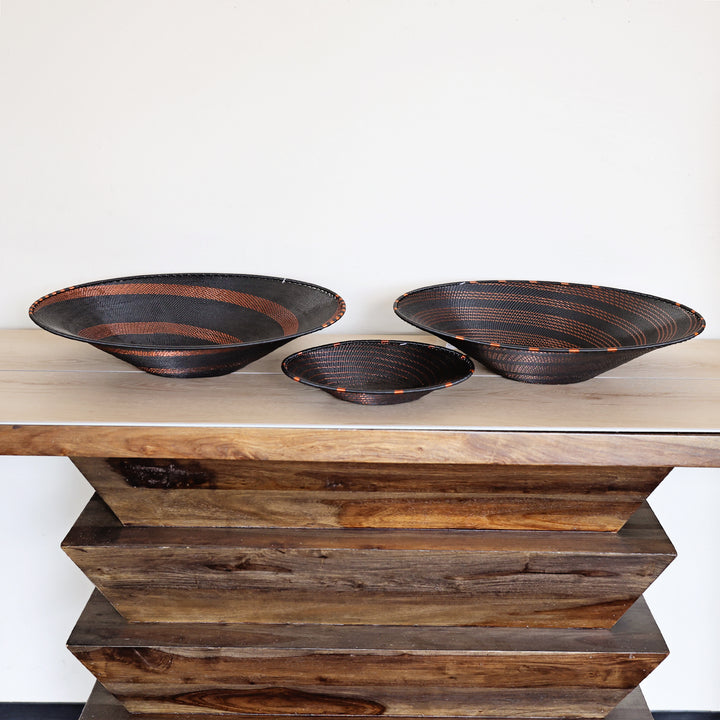 Three hand-woven dark brown and black zulu wire bowls with orange-brown accents, displayed on a rustic wooden side table against a plain white wall.