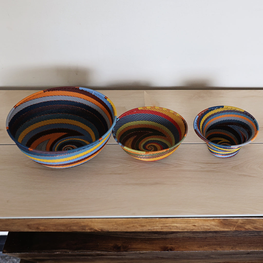 Three handwoven striped zulu bowls in different sizes displayed on a light wood tabletop against a plain white wall.