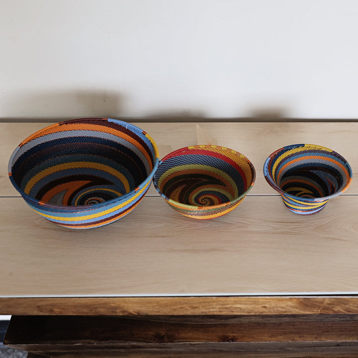 Three handwoven striped zulu bowls in different sizes displayed on a light wood tabletop against a plain white wall.