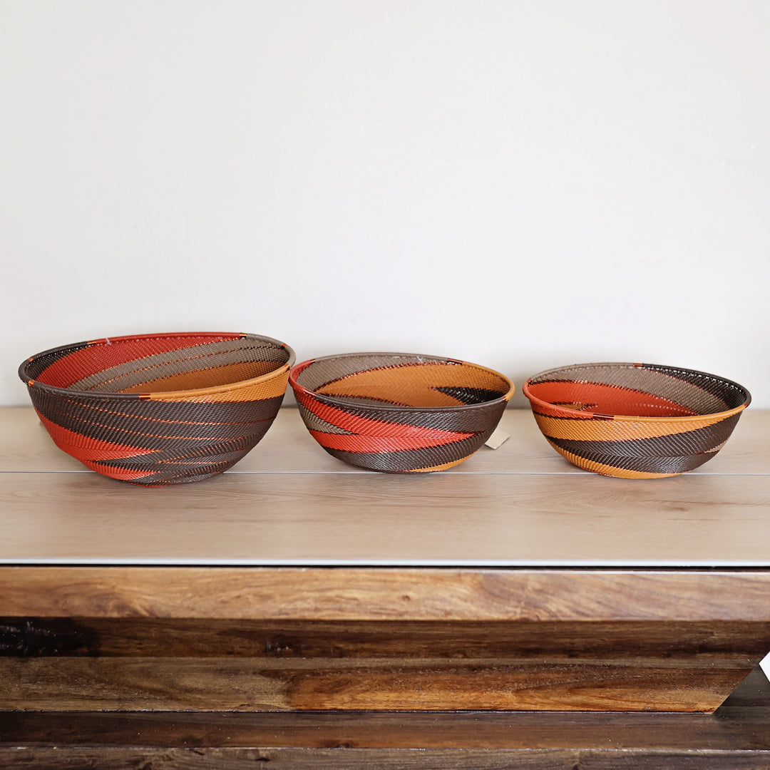 Three hand-woven Zulu baskets in different sizes, featuring a pattern of red, orange, brown, and beige stripes, displayed on a wooden surface against a plain white wall.