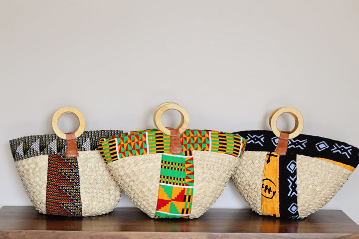 Three woven tote bags with circular wooden handles, displayed side-by-side on a dark wooden surface against a plain light gray wall. Each bag features a natural beige woven body and a vibrant Kente patterned fabric top in black, green, red, yellow, and white.