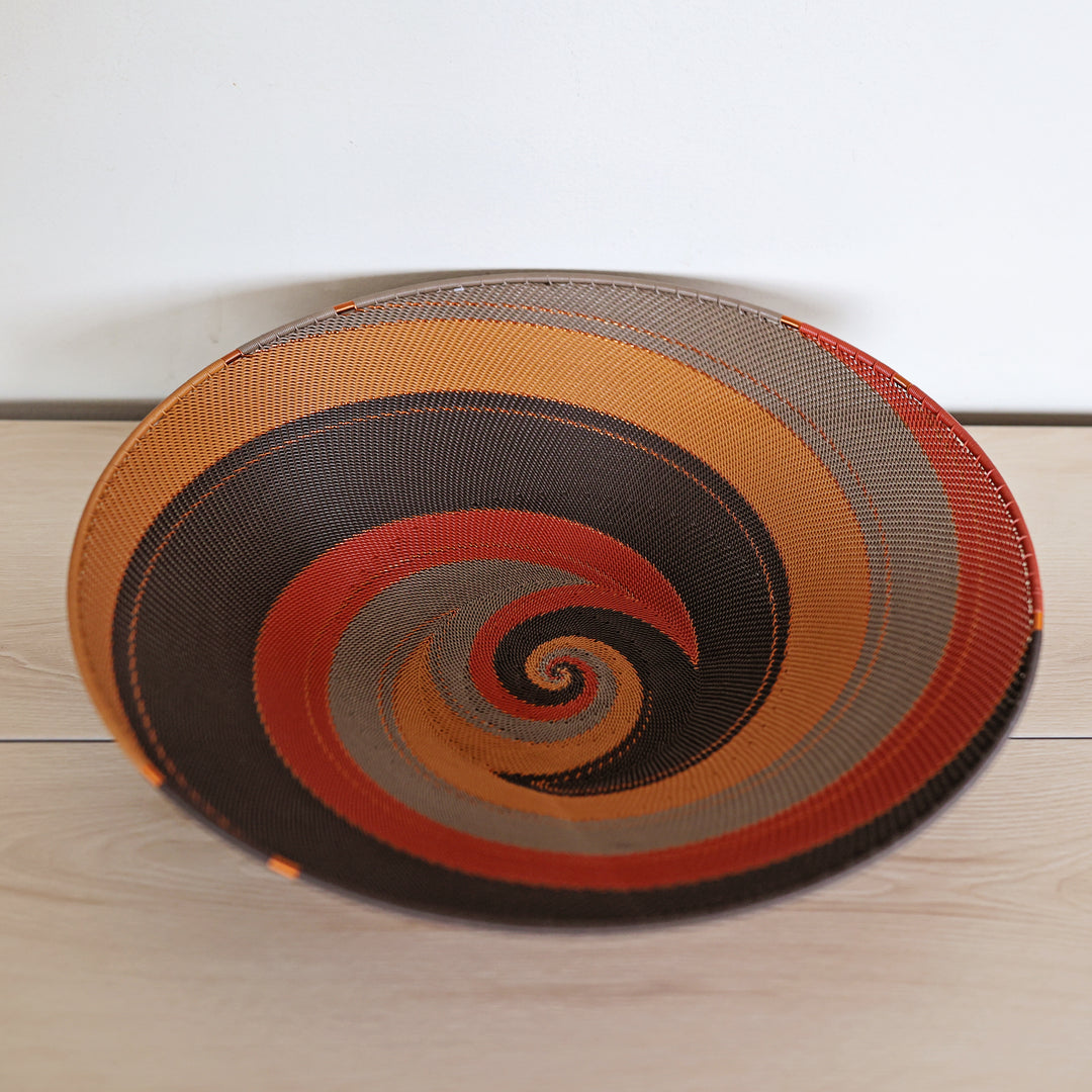 A round woven decorative Zulu bowl with a spiral pattern in orange, brown, red, and gray, resting on a light wood surface against a white wall.