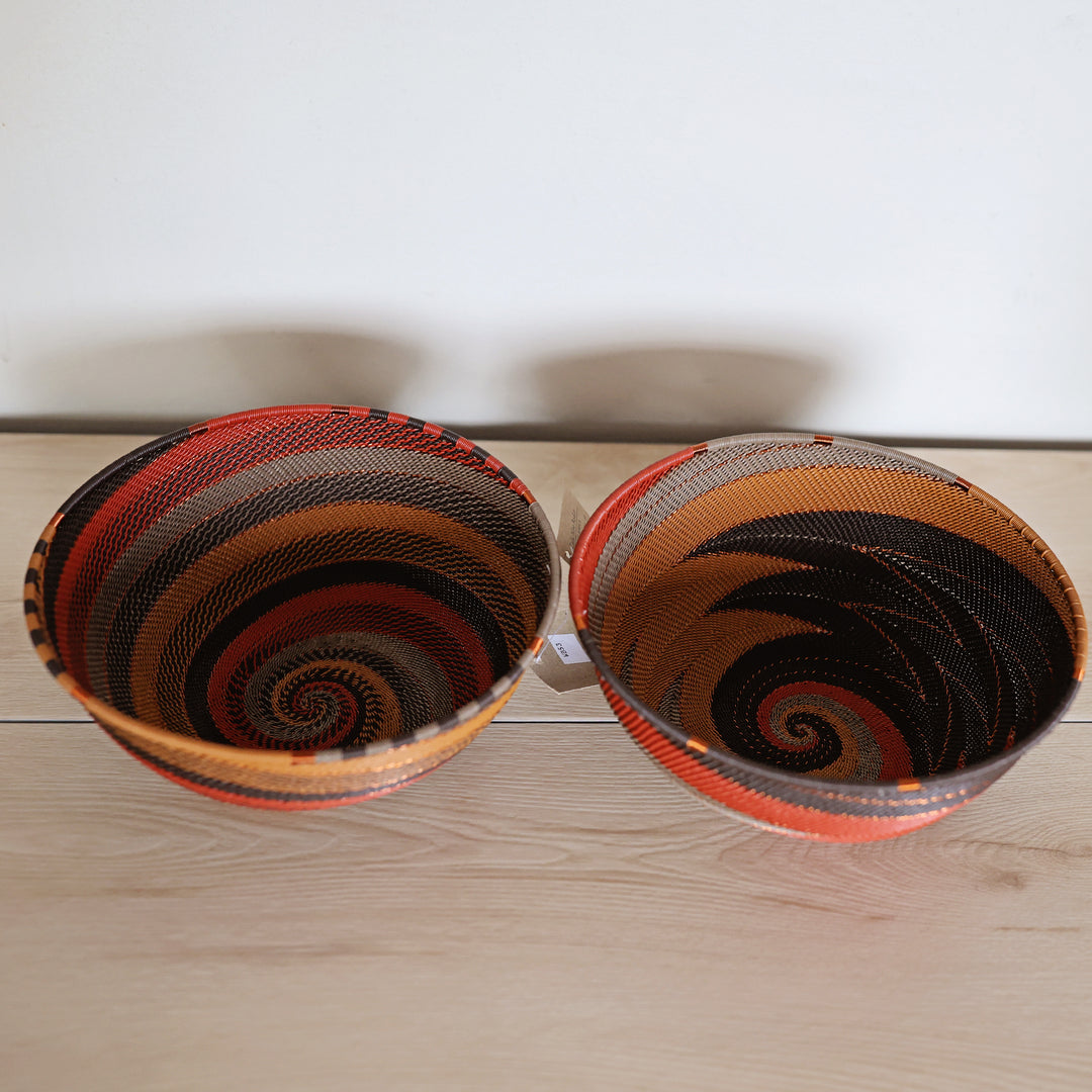 Two hand-woven Dark Earth zulu baskets with spiral patterns in orange, black, brown, and red, displayed side-by-side on a light wood surface against a white wall.