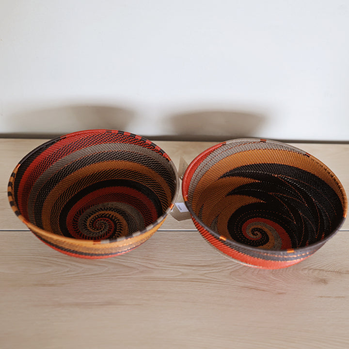 Two hand-woven Dark Earth zulu baskets with spiral patterns in orange, black, brown, and red, displayed side-by-side on a light wood surface against a white wall.