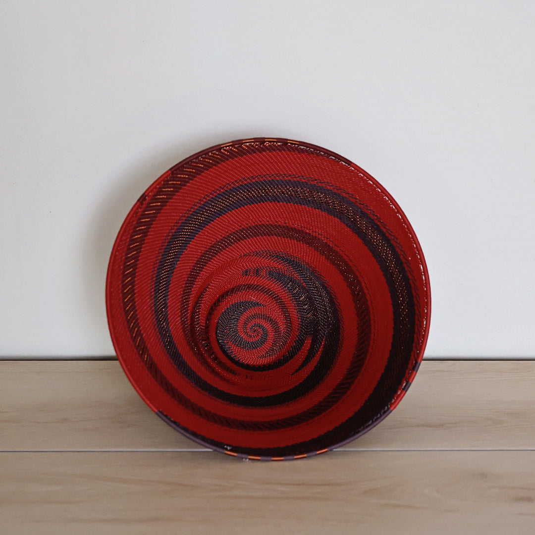 A large deep funnel hand-woven Zulu basket bowl with a bold red and black spiral pattern, standing upright on a light wood surface against a plain white wall.