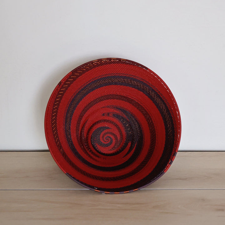 A large deep funnel hand-woven Zulu basket bowl with a bold red and black spiral pattern, standing upright on a light wood surface against a plain white wall.