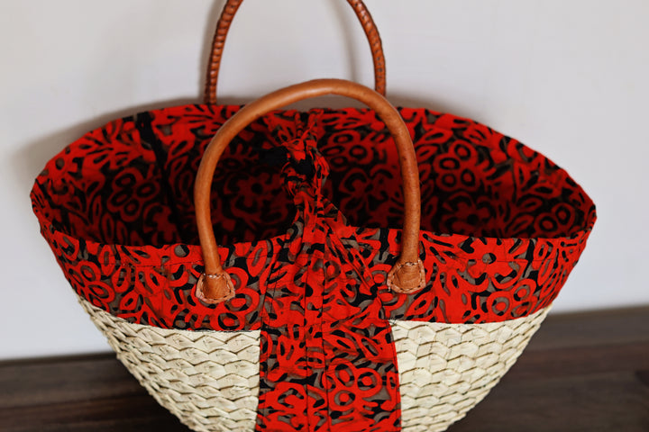 A woven straw tote bag with a red and black patterned fabric lining and brown leather handles, resting on a dark wooden surface against a plain light background.