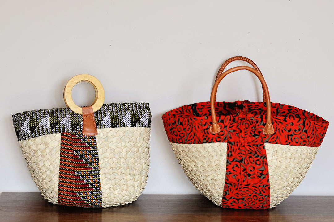 Two woven tote bags displayed side by side on a dark wooden surface against a plain light gray wall. The bag on the left has a circular light wood handle, a cream woven base, and a vertical panel with black, white, and orange geometric patterns.
