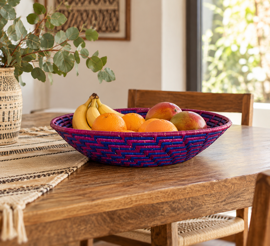 A round woven fruit bowl with a purple and blue pattern, filled with bananas, oranges, and mangoes, placed on a wooden dining table in a bright room.