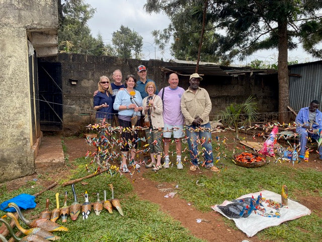 A group of seven people standing together at a workshop in Kenya , surrounded by various handmade items including colorful bead necklaces, wooden tools, and other crafts displayed on the ground. 