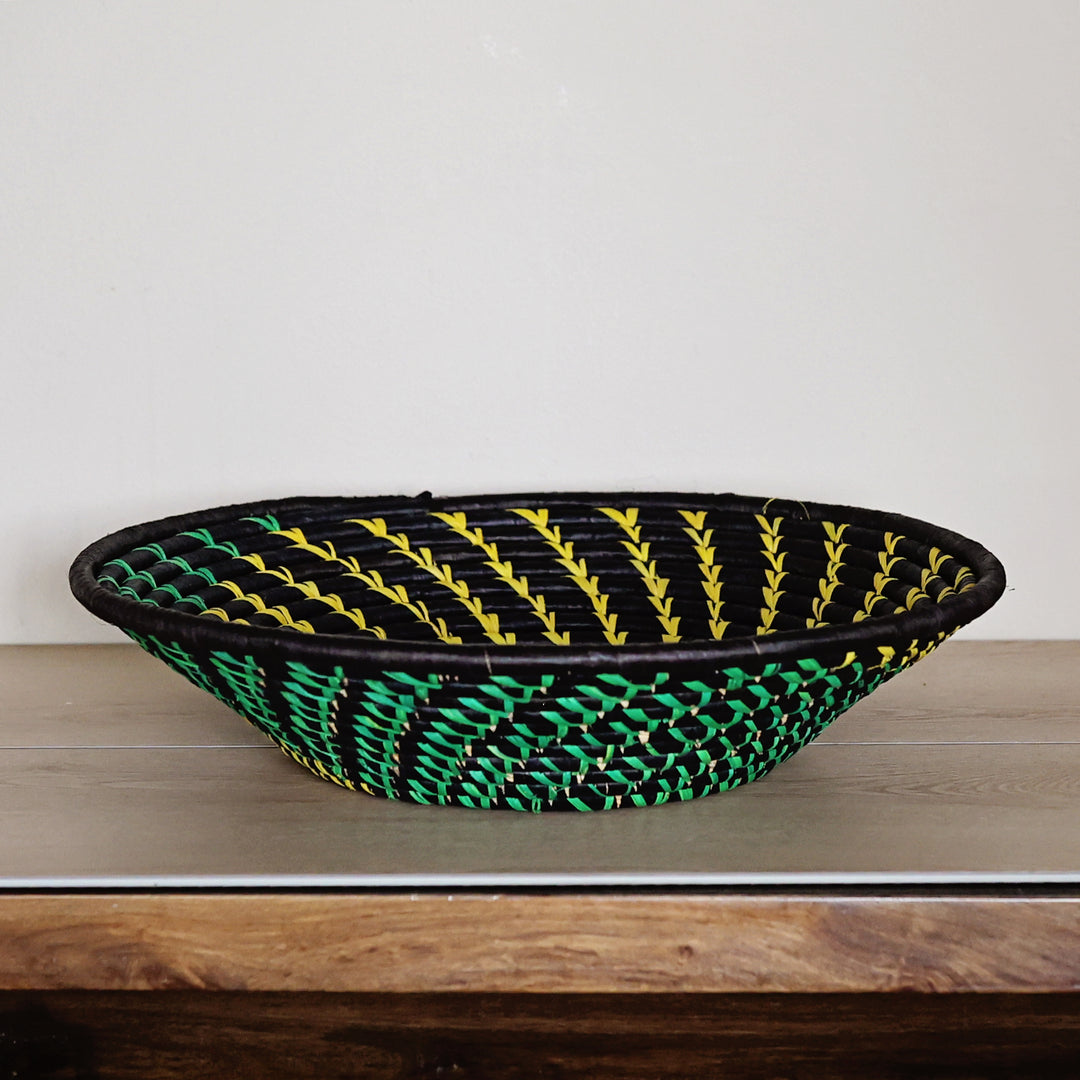 A hand-woven oval raffia basket with a black base, yellow leaf-like stripes on the upper half, and green and black woven detailing on the lower half, displayed on a wooden surface against a plain light gray wall.