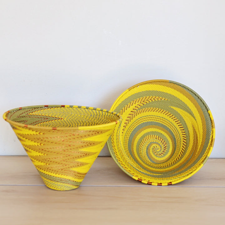 Two hand-woven papaya zulu baskets with yellow and olive green spiral patterns, displayed on a light wood surface against a plain white wall.